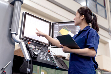 Asian woman industrial engineer in uniform touching screen display to control automated cnc machinery in industrial workshop.