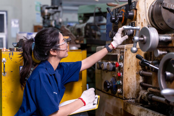 Closeup Asian woman industrial engineer in uniform checking old model lathe and drilling machine with clipboard at industrial workshop.