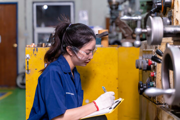 Closeup Asian woman industrial engineer in uniform checking old model lathe and drilling machine and writing a maintenance playlist on clipboard at industrial workshop.