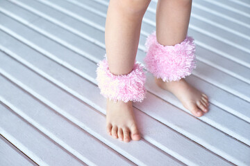 Child wearing fluffy pink ankle warmers on white wooden floor, close-up of legs and feet wearing cozy accessories.