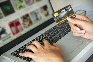 Asian man shopping online with a credit card in hand, browsing product options on a laptop—glasses and documents nearby frame a focused digital workspace vibe. Lifestyle concept.