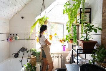 Woman in rubber gloves and apron manually washes window of house in bathroom with rag cleaner and mop inside interior with home plants. Restoring order and cleanliness, cleaning servise