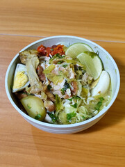 A bowl of Indonesian chicken soto with shredded chicken, sliced boiled egg, vermicelli, sliced cabbage, bean sprouts, celery, chili sauce and lime, mixed with delicious sauce. On a wooden table.