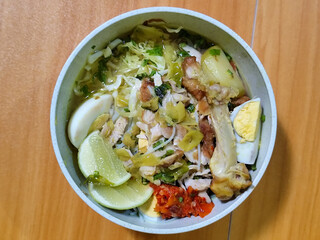 A bowl of Indonesian chicken soto with shredded chicken, sliced boiled egg, vermicelli, sliced cabbage, bean sprouts, celery, chili sauce and lime, mixed with delicious sauce. On a wooden table.