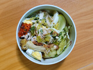 A bowl of Indonesian chicken soto with shredded chicken, sliced boiled egg, vermicelli, sliced cabbage, bean sprouts, celery, chili sauce and lime, mixed with delicious sauce. On a wooden table.