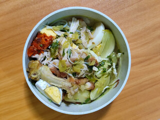 A bowl of Indonesian chicken soto with shredded chicken, sliced boiled egg, vermicelli, sliced cabbage, bean sprouts, celery, chili sauce and lime, mixed with delicious sauce. On a wooden table.