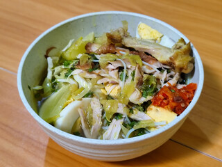 A bowl of Indonesian chicken soto with shredded chicken, sliced boiled egg, vermicelli, sliced cabbage, bean sprouts, celery, chili sauce and lime, mixed with delicious sauce. On a wooden table.