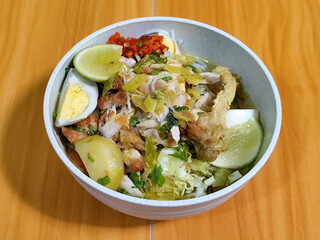 A bowl of Indonesian chicken soto with shredded chicken, sliced boiled egg, vermicelli, sliced cabbage, bean sprouts, celery, chili sauce and lime, mixed with delicious sauce. On a wooden table.