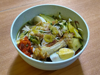A bowl of Indonesian chicken soto with shredded chicken, sliced boiled egg, vermicelli, sliced cabbage, bean sprouts, celery, chili sauce and lime, mixed with delicious sauce. On a wooden table.