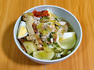 A bowl of Indonesian chicken soto with shredded chicken, sliced boiled egg, vermicelli, sliced cabbage, bean sprouts, celery, chili sauce and lime, mixed with delicious sauce. On a wooden table.