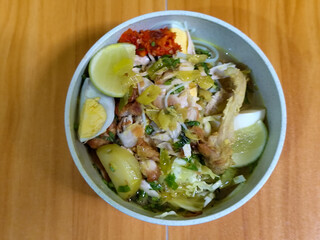 A bowl of Indonesian chicken soto with shredded chicken, sliced boiled egg, vermicelli, sliced cabbage, bean sprouts, celery, chili sauce and lime, mixed with delicious sauce. On a wooden table.