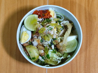 A bowl of Indonesian chicken soto with shredded chicken, sliced boiled egg, vermicelli, sliced cabbage, bean sprouts, celery, chili sauce and lime, mixed with delicious sauce. On a wooden table.