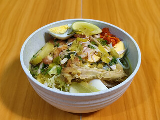 A bowl of Indonesian chicken soto with shredded chicken, sliced boiled egg, vermicelli, sliced cabbage, bean sprouts, celery, chili sauce and lime, mixed with delicious sauce. On a wooden table.