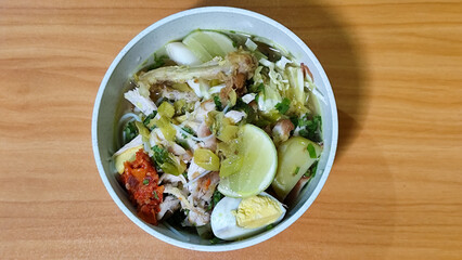 A bowl of Indonesian chicken soto with shredded chicken, sliced boiled egg, vermicelli, sliced cabbage, bean sprouts, celery, chili sauce and lime, mixed with delicious sauce. On a wooden table.