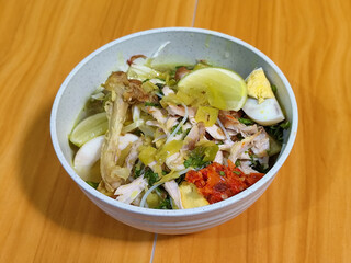 A bowl of Indonesian chicken soto with shredded chicken, sliced boiled egg, vermicelli, sliced cabbage, bean sprouts, celery, chili sauce and lime, mixed with delicious sauce. On a wooden table.