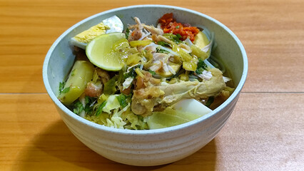 A bowl of Indonesian chicken soto with shredded chicken, sliced boiled egg, vermicelli, sliced cabbage, bean sprouts, celery, chili sauce and lime, mixed with delicious sauce. On a wooden table.