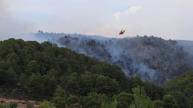 Helicoptero no reconocible con Bambi Bucket se aproxima al fuego en incendio forestal del parque natural de la Font Roja en julio de 2025, Ibi, Espa&ntilde;a
