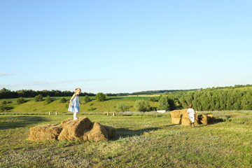 Two children enjoy a delightful summer day, climbing on stacked hay bales in a lush green field...