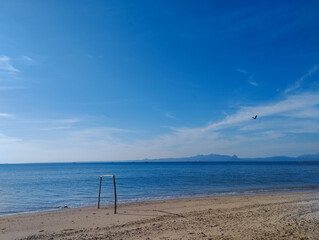 A bird soars through a clear blue sky above a calm beach scene. A lone wooden frame stands near the shoreline, facing the serene ocean. Distant mountains fade into the horizon, completing this peacefu