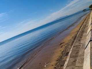 A calm shoreline view featuring a stone seawall on the right and gentle ocean waves lapping onto the sandy beach under a vivid blue sky. Distant landmasses stretch across the horizon, offering a tranq