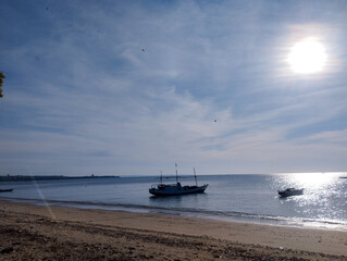 A peaceful coastal scene with traditional boats floating on calm waters under a bright morning sun. The sunlight creates shimmering reflections on the sea, while the beach remains quiet and undisturbe