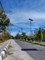 A quiet asphalt road with a pedestrian sidewalk and solar-powered street lamps stretches through a semi-urban area, surrounded by trees and vegetation under a vivid blue sky with scattered clouds