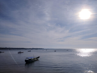 A traditional fishing boat floats calmly on shimmering sea waters beneath a bright morning sun. Sunlight reflects across the peaceful ocean, casting a silver glow on the horizon. Other boats scatter i
