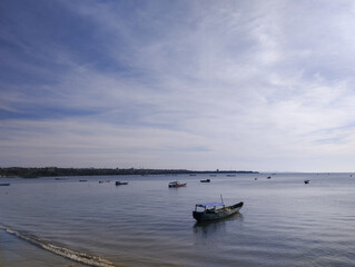 A traditional wooden boat floats quietly near the shore under a bright and lightly clouded sky. Gentle ripples meet the coastline, while several boats are scattered across the calm sea. In the distanc