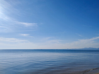 A wide-angle view of a tranquil ocean meeting the horizon under a clear, vibrant blue sky. Gentle ripples form on the calm sea surface, with distant mountains creating a peaceful backdrop. The minimal
