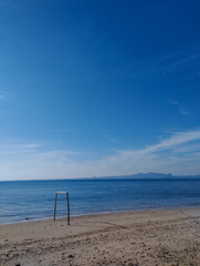A solitary wooden frame stands on a quiet sandy beach facing the calm sea, under a vast clear blue sky. The distant mountains create a serene backdrop, while the soft shoreline and tranquil water evok