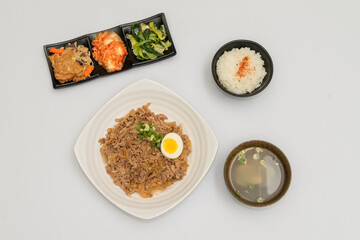 A savory beef rice bowl served as a set meal, featuring tender slices of beef simmered in soy-based sauce atop steamed white rice. The dish is accompanied by a variety of side vegetables and a light