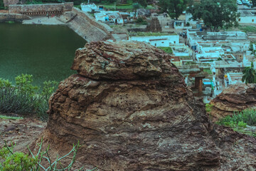 Rugged rock formation overlooks an Indian village and tranquil pond, showcasing layered textures...