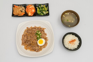 A savory beef rice bowl served as a set meal, featuring tender slices of beef simmered in soy-based sauce atop steamed white rice. The dish is accompanied by a variety of side vegetables and a light