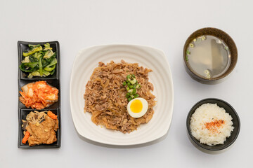 A savory beef rice bowl served as a set meal, featuring tender slices of beef simmered in soy-based sauce atop steamed white rice. The dish is accompanied by a variety of side vegetables and a light