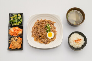 A savory beef rice bowl served as a set meal, featuring tender slices of beef simmered in soy-based sauce atop steamed white rice. The dish is accompanied by a variety of side vegetables and a light