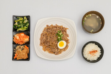 A savory beef rice bowl served as a set meal, featuring tender slices of beef simmered in soy-based sauce atop steamed white rice. The dish is accompanied by a variety of side vegetables and a light