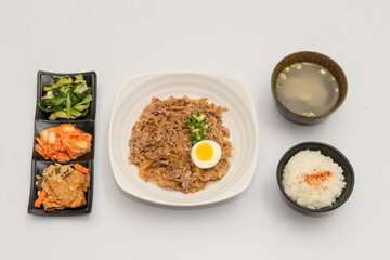 A savory beef rice bowl served as a set meal, featuring tender slices of beef simmered in soy-based sauce atop steamed white rice. The dish is accompanied by a variety of side vegetables and a light