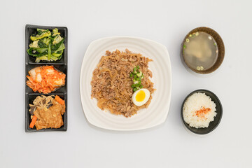 A savory beef rice bowl served as a set meal, featuring tender slices of beef simmered in soy-based sauce atop steamed white rice. The dish is accompanied by a variety of side vegetables and a light