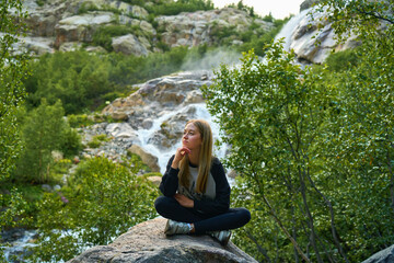 A tourist girl is sitting on a rock by a waterfall