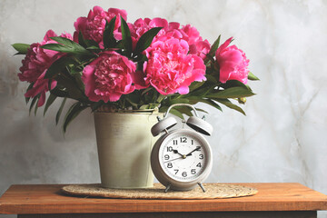 Pink peonies in a metal vase and a gray alarm clock standing on a wooden table