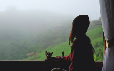 A woman with a cat sitting on bed while looking at a beautiful nature view on foggy morning