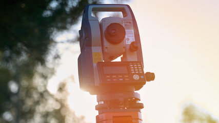 A surveying instrument is standing in the sunlight. The instrument is a total station, which is used to measure angles and distances. The instrument is positioned on a tripod