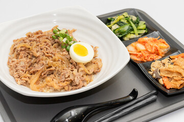 A savory beef rice bowl served as a set meal, featuring tender slices of beef simmered in soy-based sauce atop steamed white rice. The dish is accompanied by a variety of side vegetables and a light