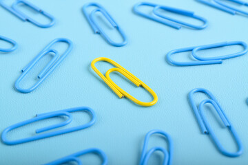 Yellow paper clip among blue ones on light blue background, top view. Difference, individuality,...