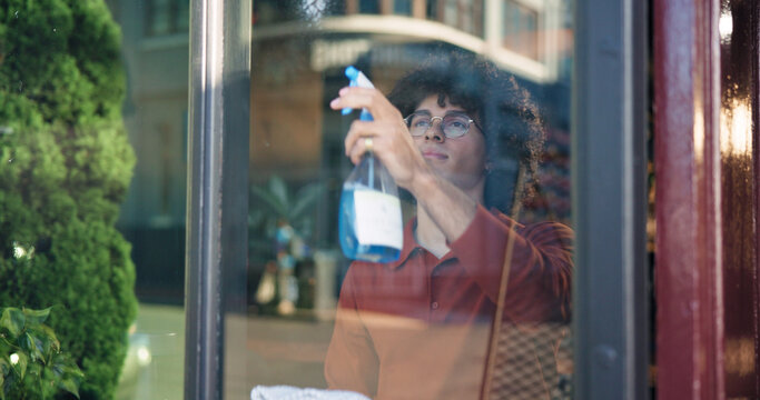 Man, owner and cleaning with window in cafe for detergence, disinfectant or preparation. Male person, barista or bottle with glass, spray or chemical for dirt, bacteria or germ removal in coffee shop