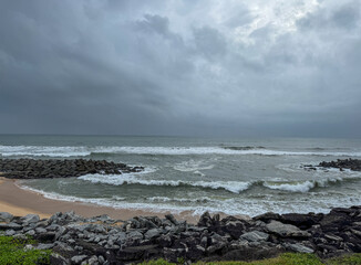 View of a beach with waves ,sand ,rocks ,greenery and blue sky.