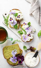 Breakfast cream cheese toast with pansies flowers and spinach bread on plate, on white marble background.