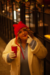 Plus size woman walks around the city during the Christmas of the week with a hot drink in a glass against the background of flickering light bulbs