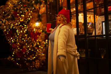 Plus size woman walks around the city during the Christmas of the week with a hot drink in a glass against the background of flickering light bulbs