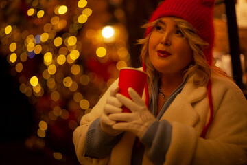 Plus size woman walks around the city during the Christmas of the week with a hot drink in a glass against the background of flickering light bulbs